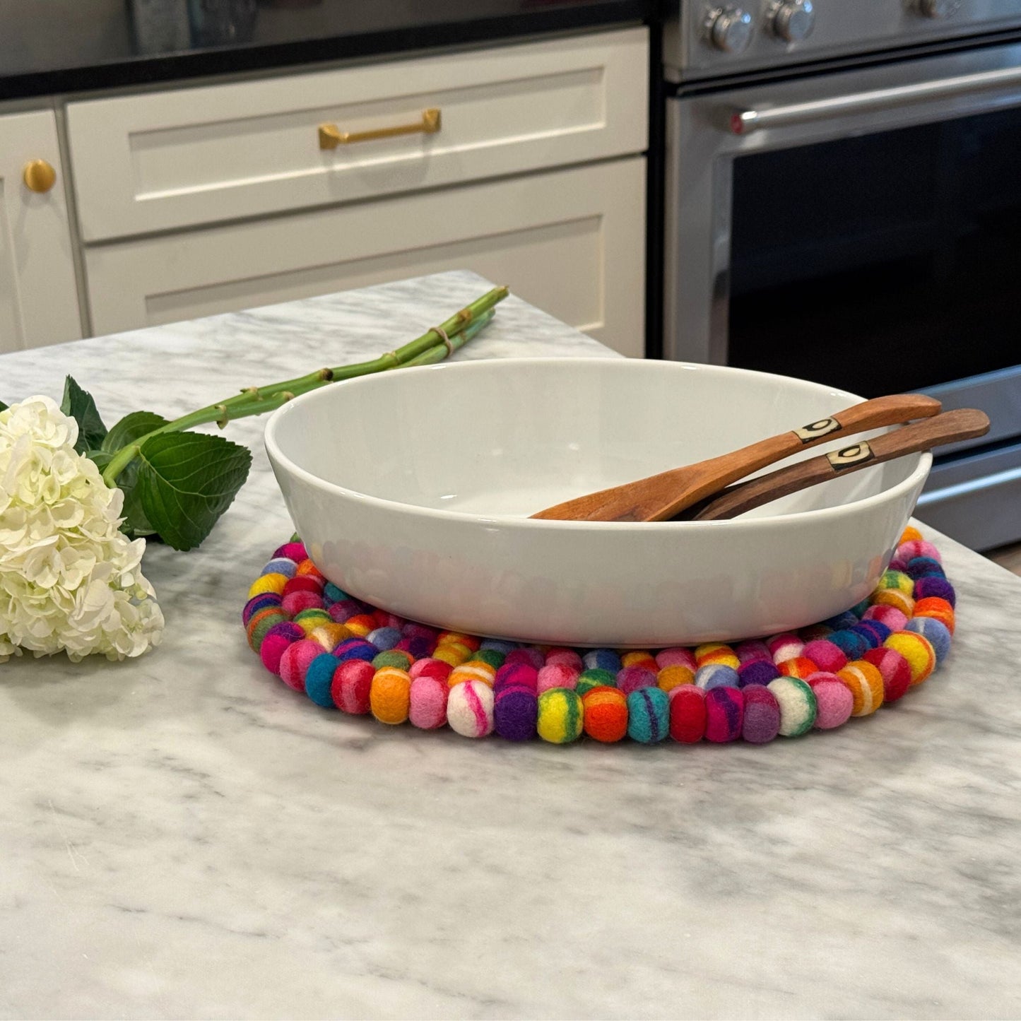 White bowl with wooden spoons on a  colorful rainbow large oval felt ball  trivet on a kitchen counter.
