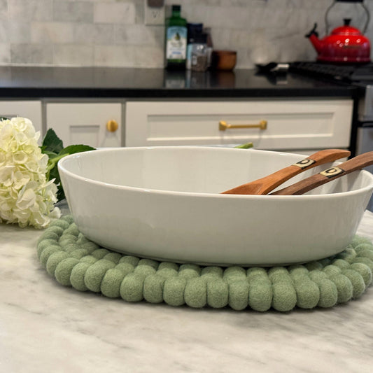 White casserole dish with wooden spoons on a sage green felt ball oval  trivet in a kitchen setting