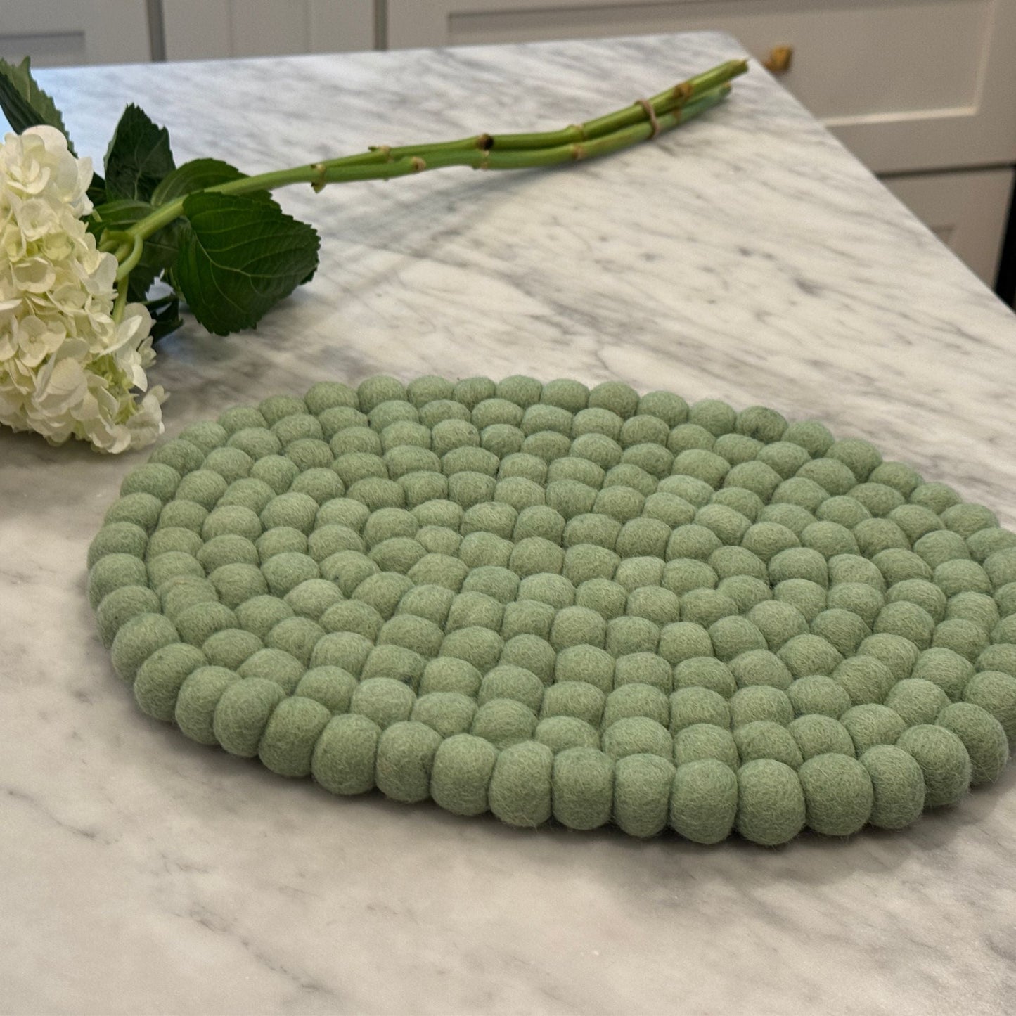 Green felt ball oval trivet on a marble surface with flowers.