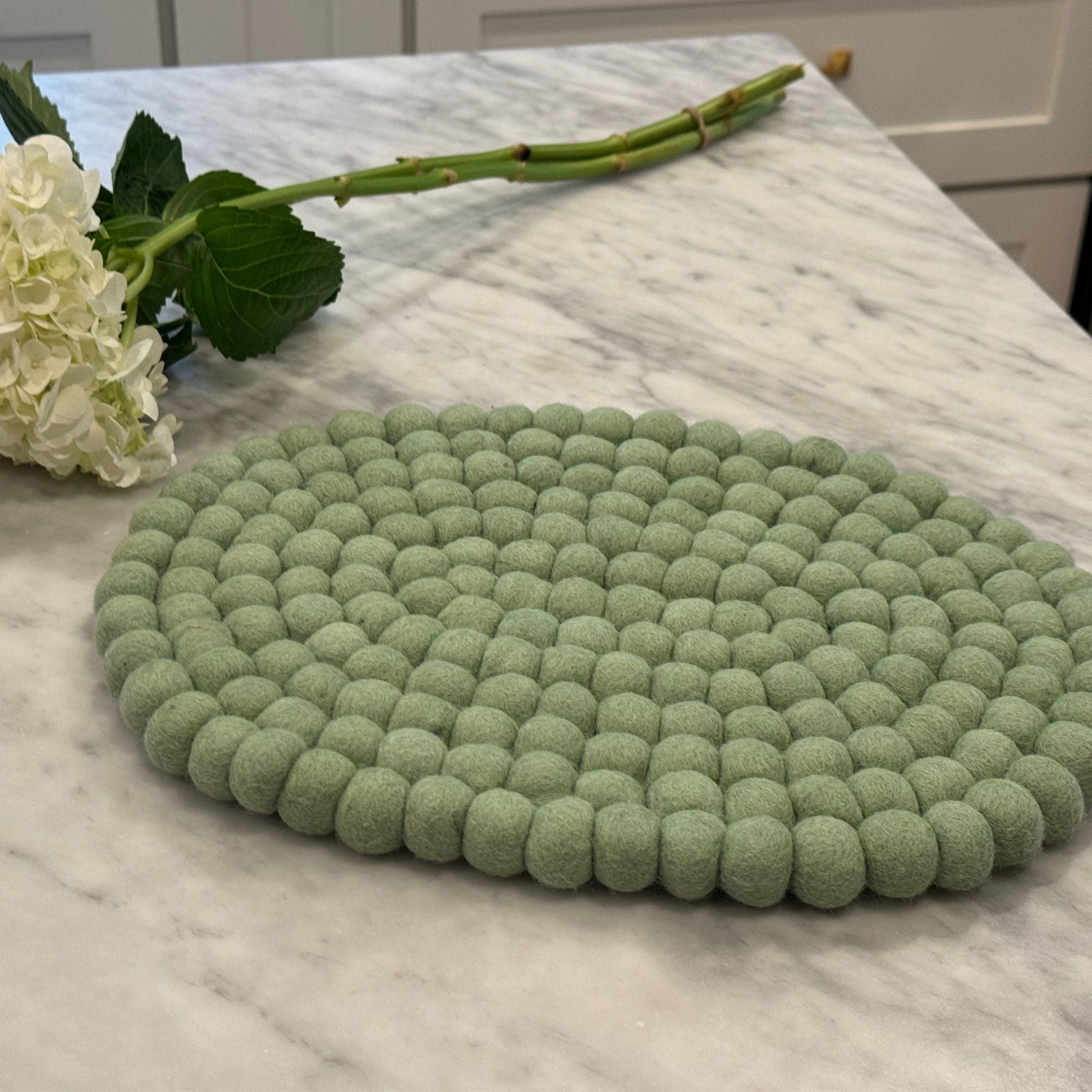 Green felt ball oval trivet on a marble surface with flowers.