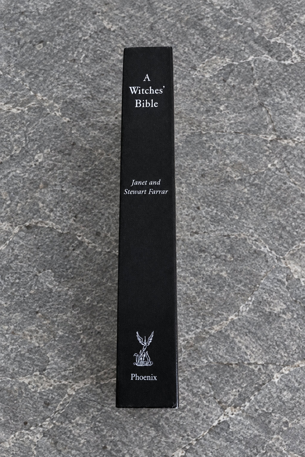 A tall, narrow black book spine standing upright on a light wood-grained surface. The spine reads 'A Witches' Bible' with 'Janet and Stewart Farrar' and the 'Phoenix' publisher logo near the bottom.