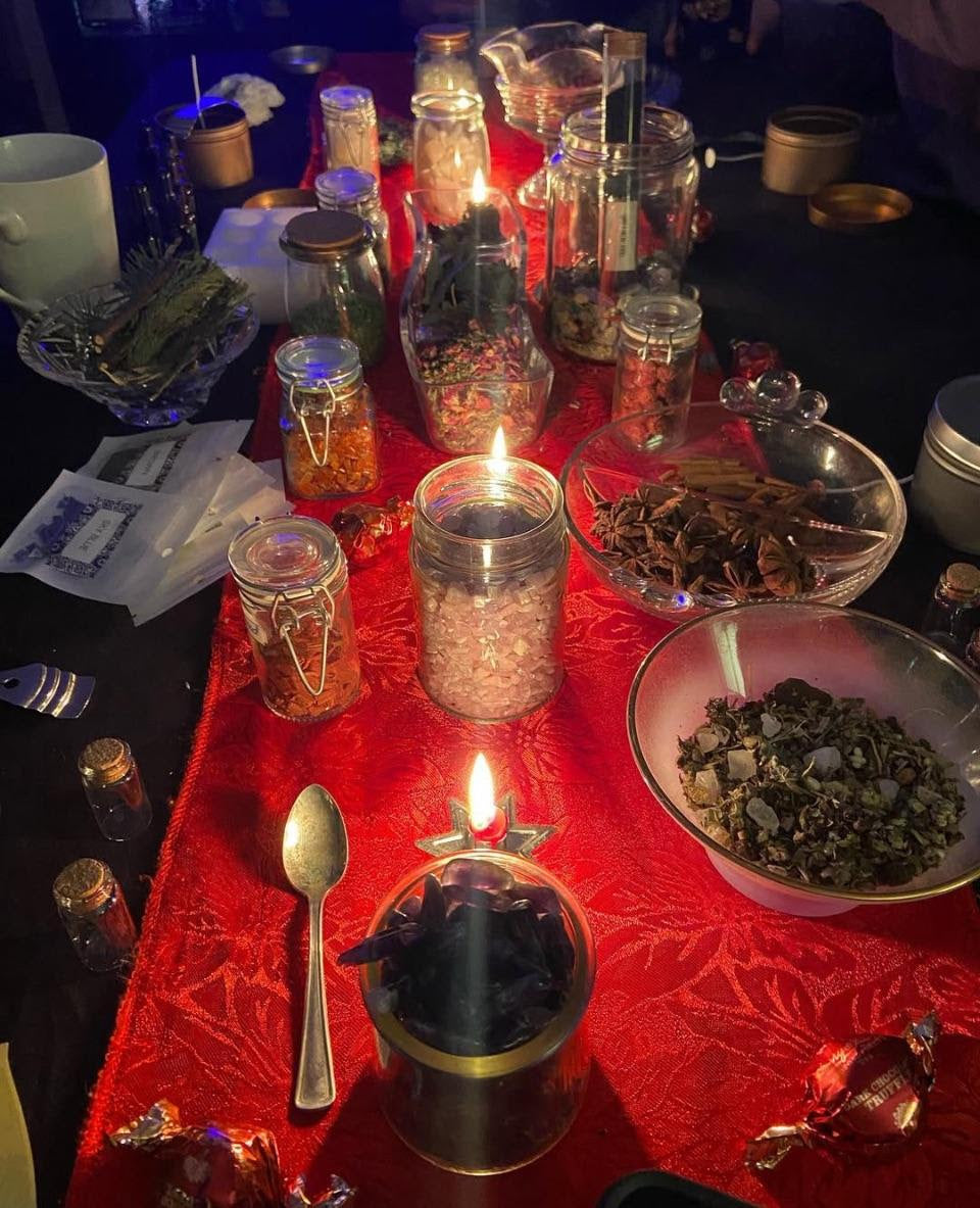 Table with various items used in ritual magic and baths, including jars, a bowl, and a lit candle on a red cloth.