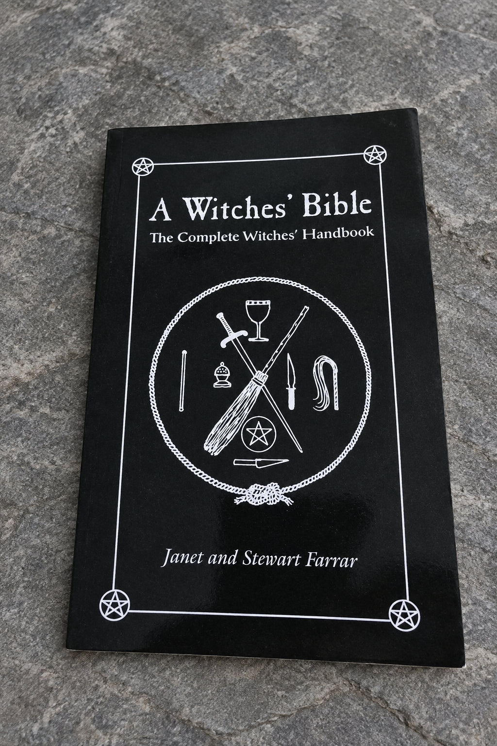 The front cover of the book 'A Witches' Bible: The Complete Witches' Handbook' by Janet and Stewart Farrar, featuring a black background with white text and occult symbols, photographed on a light wood surface.